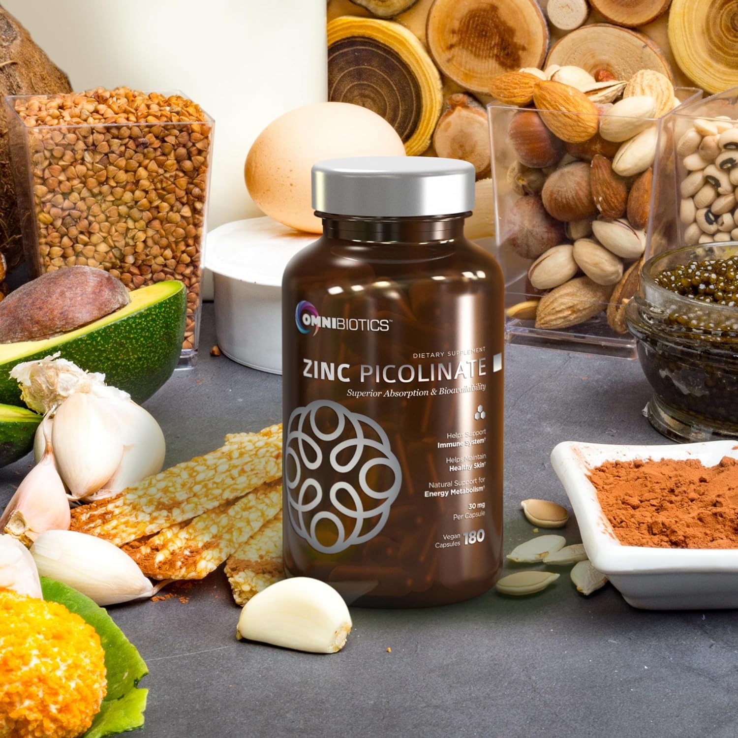 OmniBiotics Zinc Picolinate 30 mg (180 vegetarian capsules) supplement bottle on a counter surrounded by zinc-rich foods—nuts, seeds, avocado, garlic, legumes, and cocoa powder.